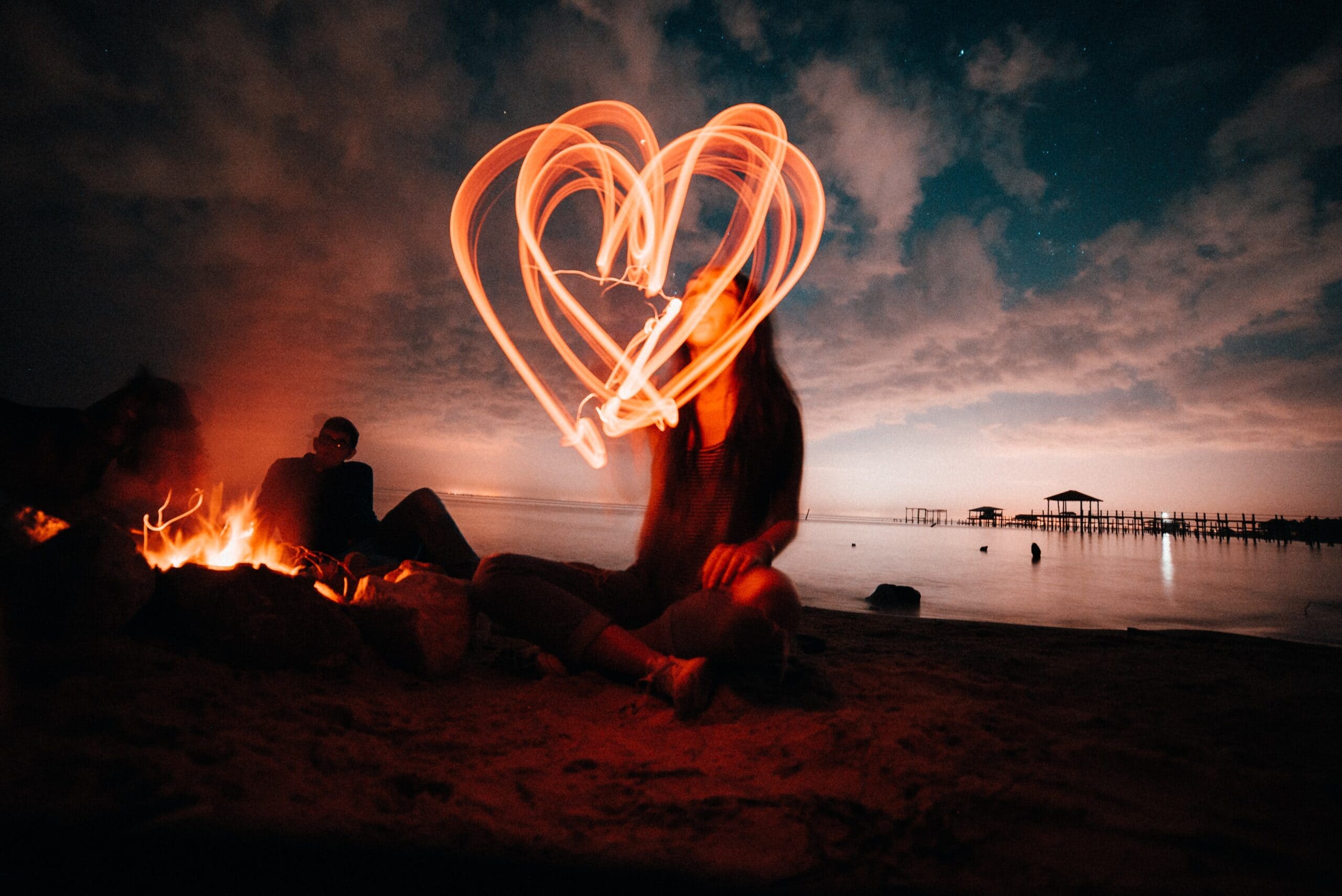 A long exposure red heart drawn n front of a figure next to a campfire at sunset.